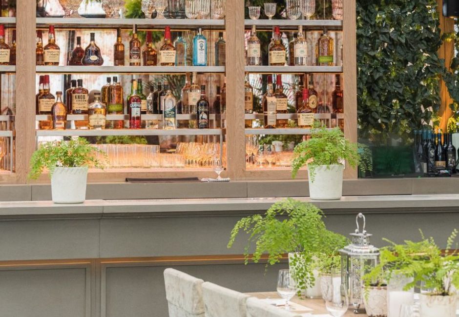Outdoor kitchen with a gray wooden bar area with the liquor bottles encased in a cabinet with glass doors; a natural wood table is surrounded by linen-covered chairs; ferns are featured hanging from above as well as potted on the bar and the table