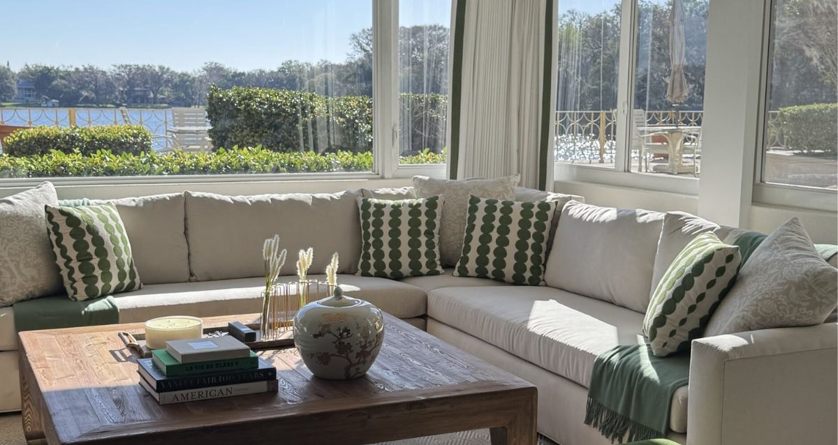 Living room setting with white couches and forest green and white pillows, surrounded by open windows that bring in natural light