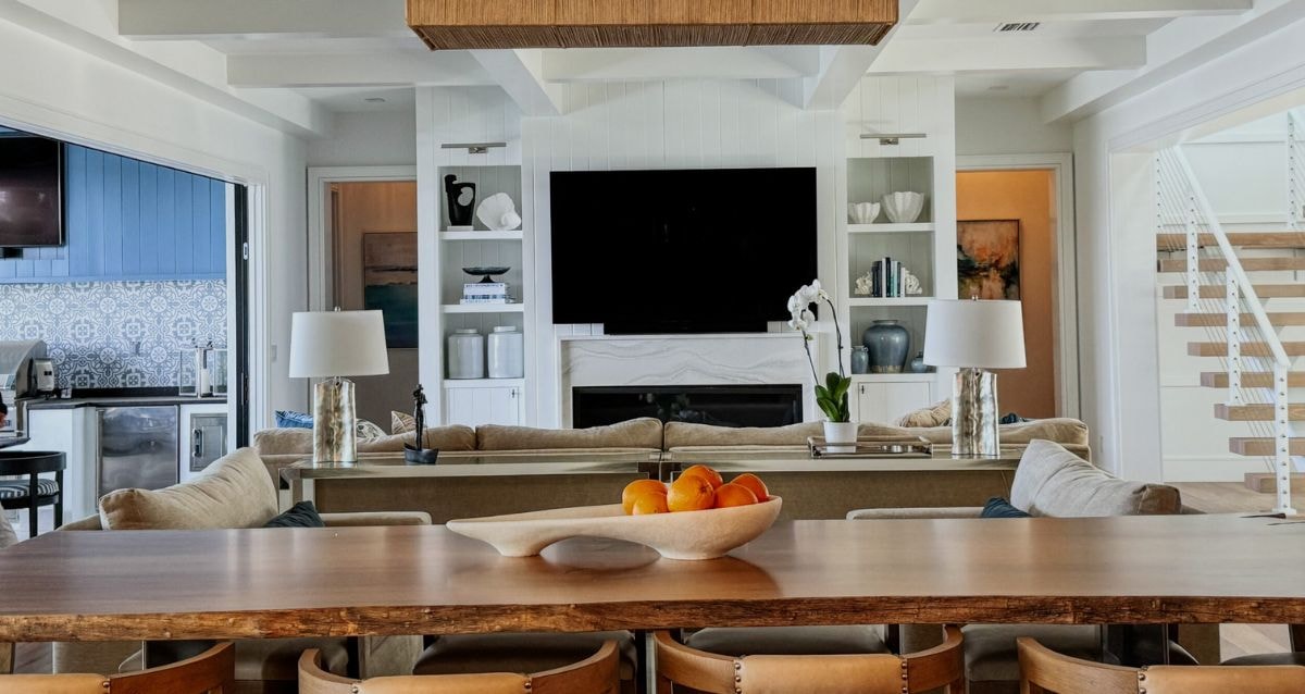 Open plan living room with wooden dining table, neutral couches, white television unit, floating stairs leading up and a kitchen space with blue color accents
